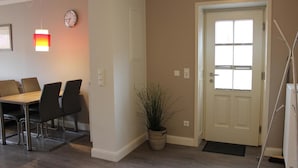 A dining area with a wooden table and chairs, a potted plant, a door with a window, and a wall-mounted clock. - Altstadt Villa Wohnung 3 Memmert (Borkum)