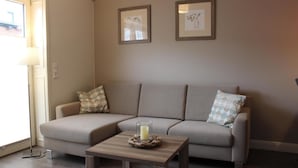 A living room with a beige sectional sofa, two framed artworks on the wall, a small wooden coffee table with a candle, and a floor lamp. - Altstadt Villa Wohnung 3 Memmert (Borkum)
