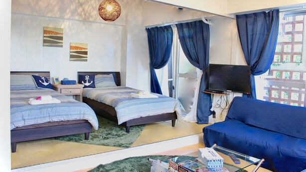 A hotel room with two beds, a sofa, a TV, and a large window with curtains. - Holiday Homes TSUBOYA 4th (Naha)