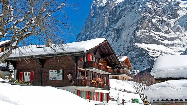 Een houten chalet met meerdere balkons, omgeven door besneeuwde bergen en een helderblauwe lucht.