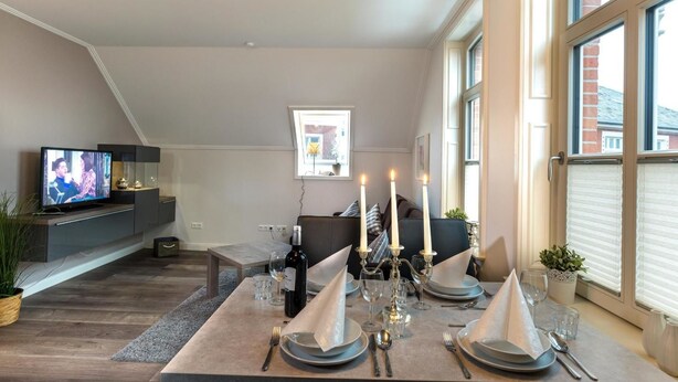 Un salon moderne doté d’un téléviseur à écran plat, d’un coin repas aménagé pour quatre personnes avec des bougies, et d’une grande fenêtre équipée de stores.