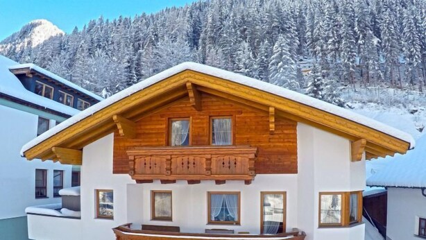 Eine zweistöckige Chalet mit Holzbalkon und einem mit Schnee bedeckten Dach, eingebettet in eine bewaldete Berglandschaft.