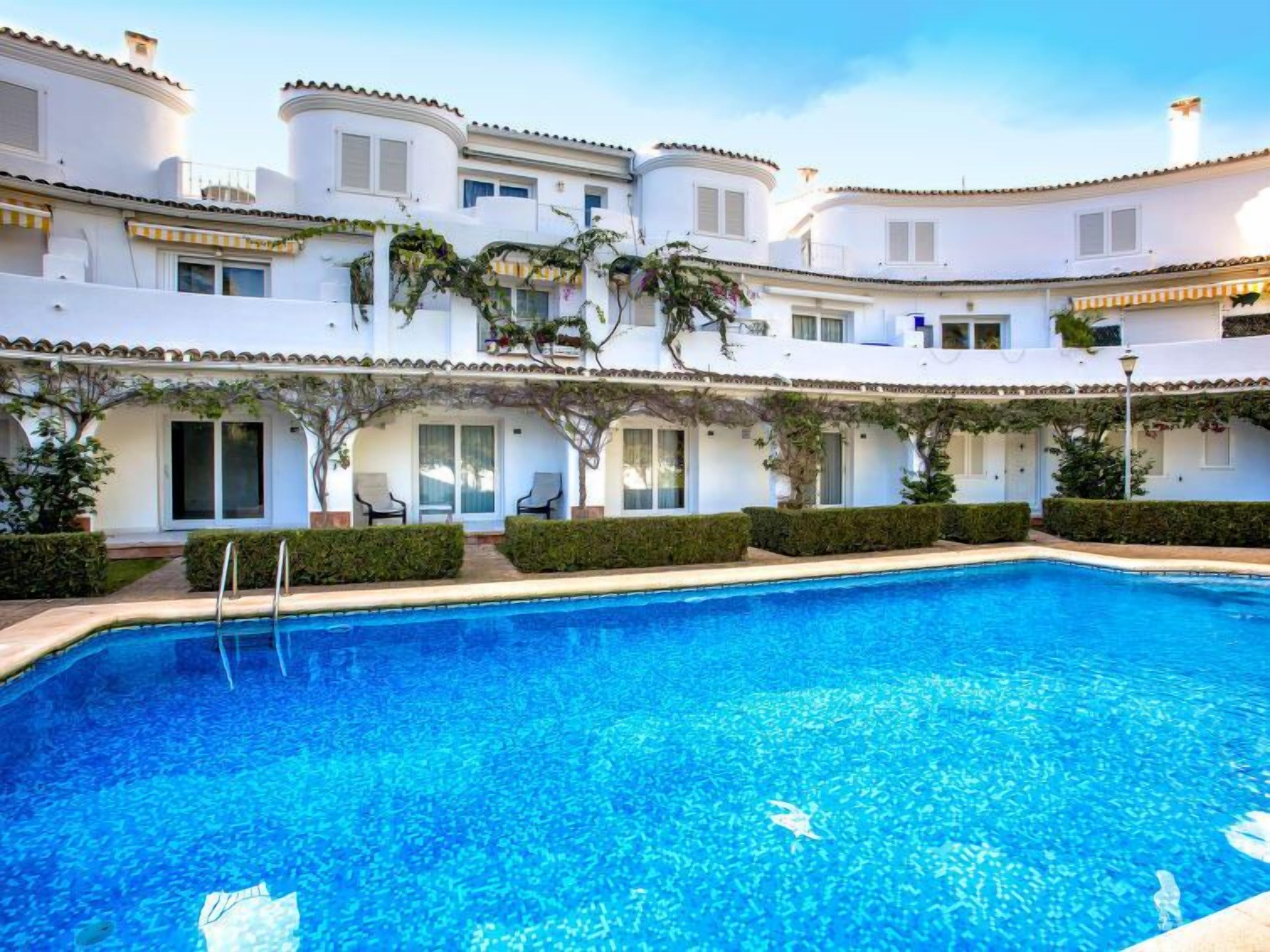 A swimming pool with a clear blue water surface, surrounded by white buildings with balconies and covered outdoor seating areas.
