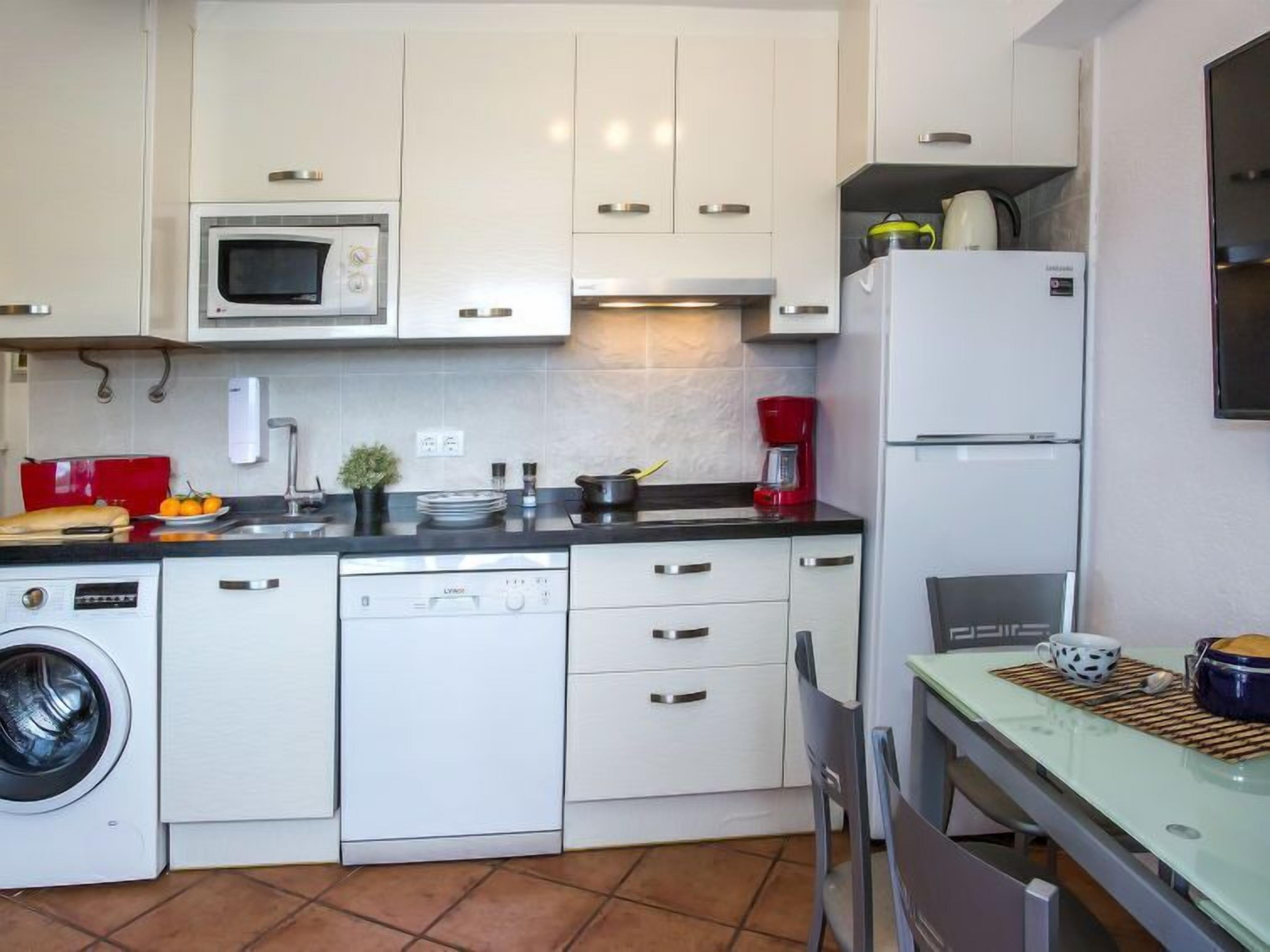 Eine kompakte Küche mit weißen Schränken, einer Mikrowelle, einem Geschirrspüler, einer Kaffeemaschine und einem Kühlschrank.