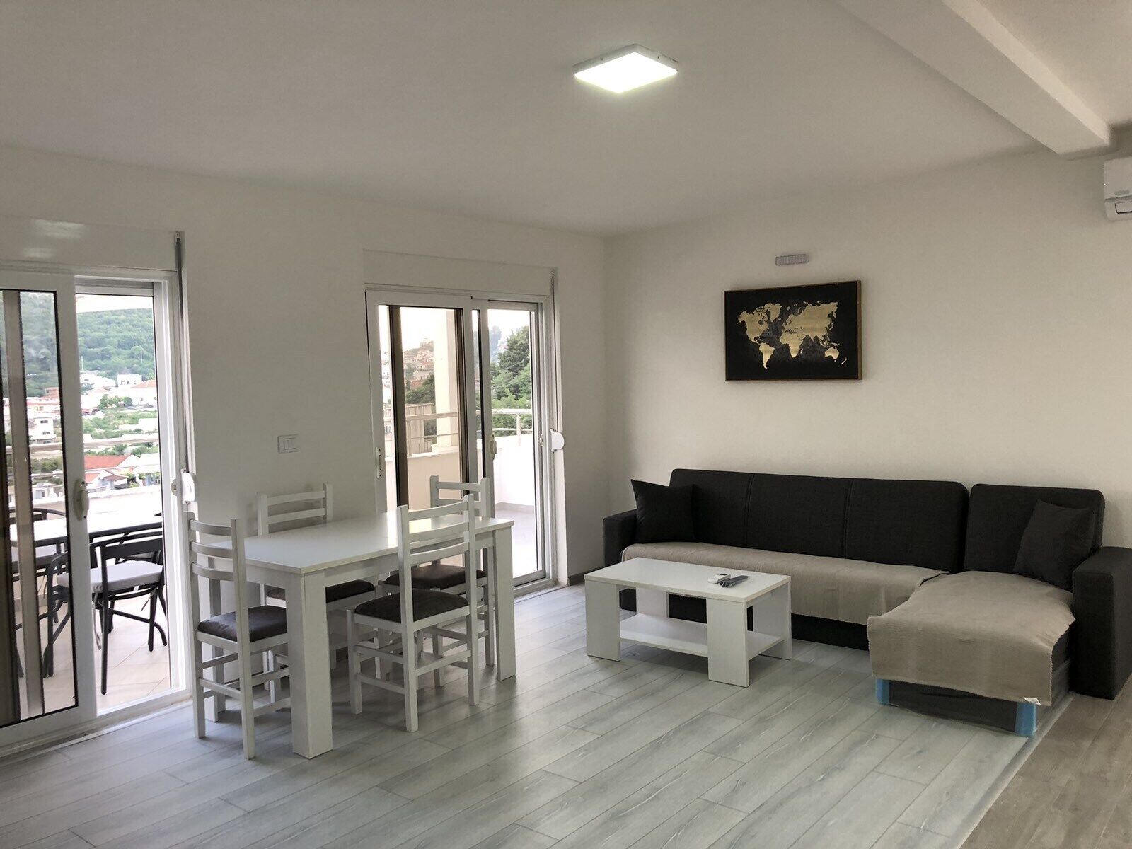 A modern living room with a white dining table and chairs, a grey sofa, and a small white coffee table. There is a large window with a view of buildings and greenery outside.