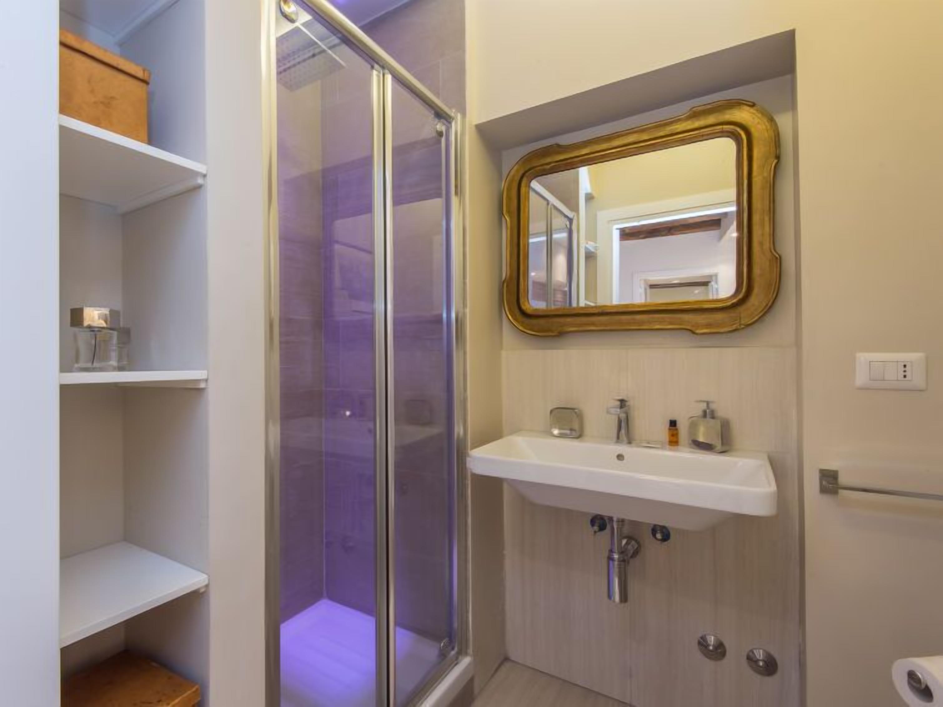 Un bagno con box doccia in vetro, un lavandino bianco con specchio dorato e scaffali in legno.