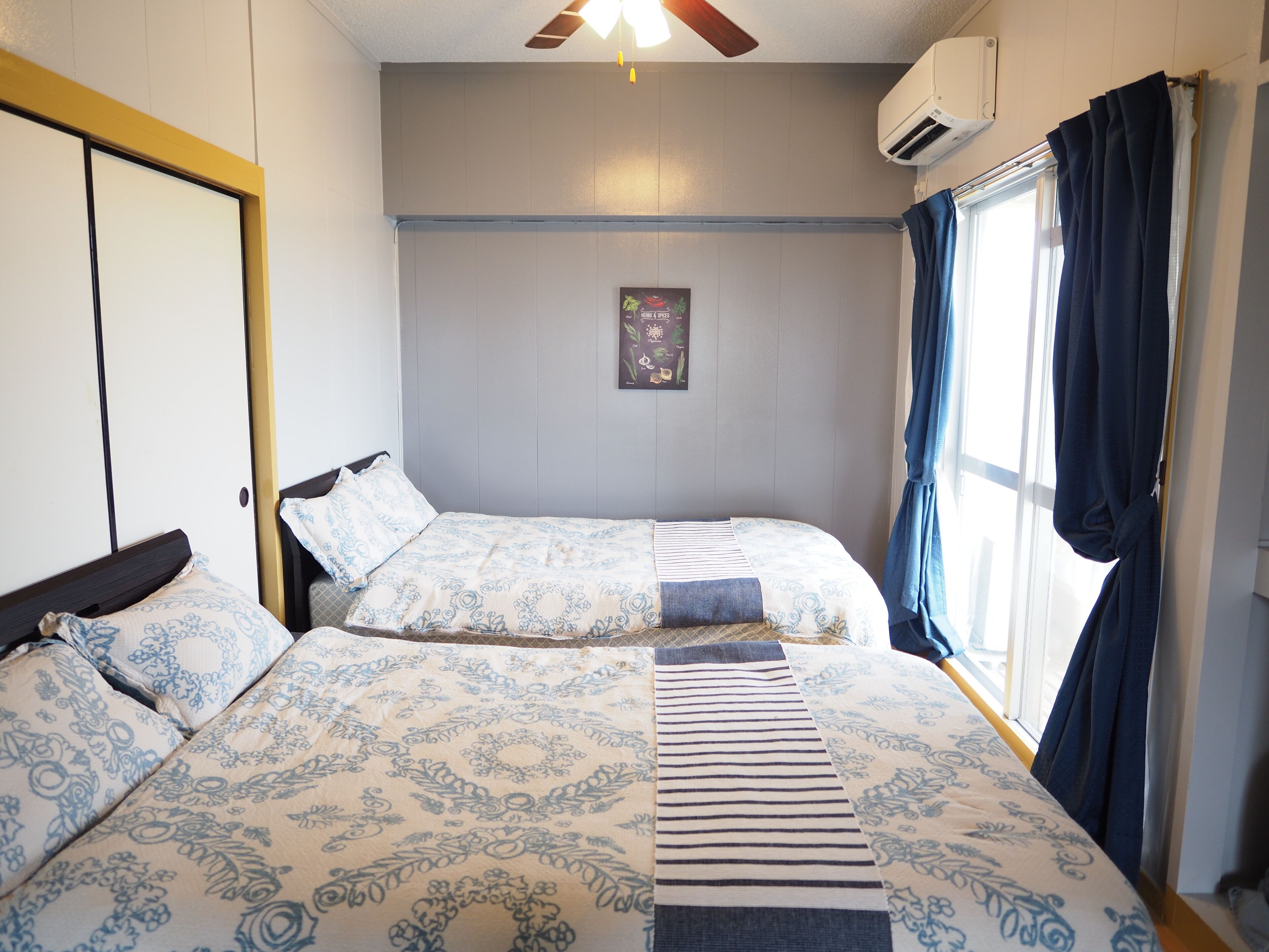 A room with two beds, a ceiling fan, an air conditioning unit, and a window with curtains.