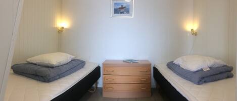 A small, compact room with two single beds, a small wooden dresser, and two wall-mounted lights.