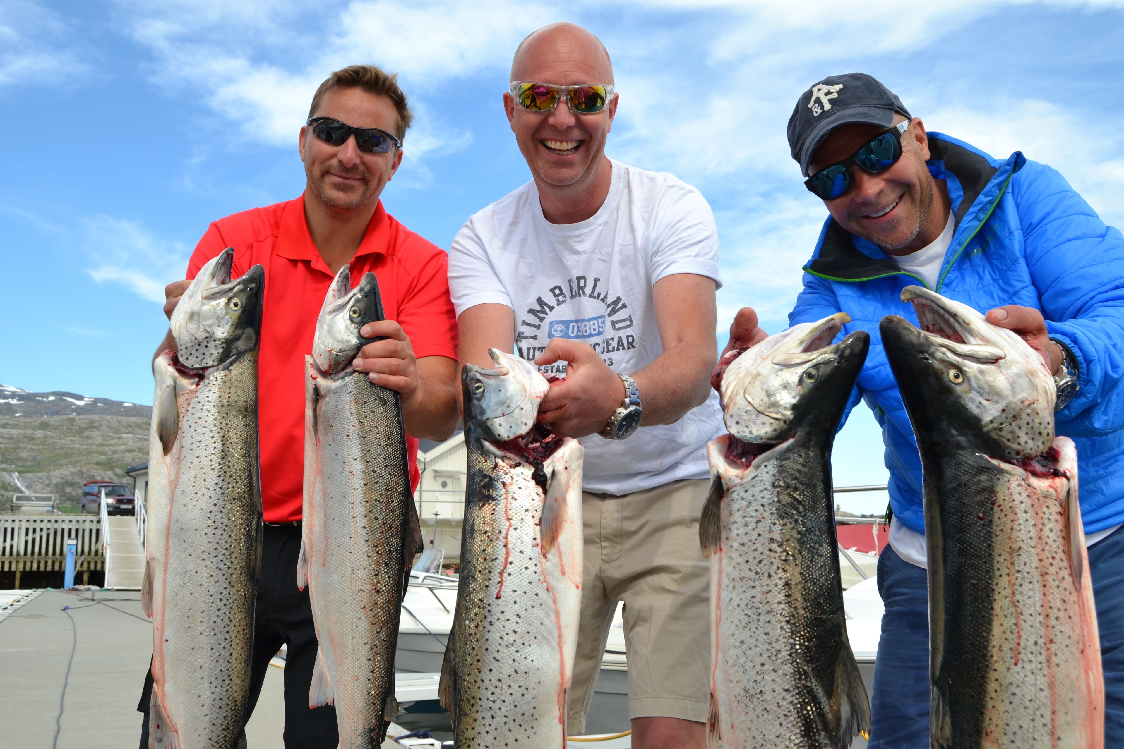 Tres personas sostienen peces en un muelle, con un bote al fondo.