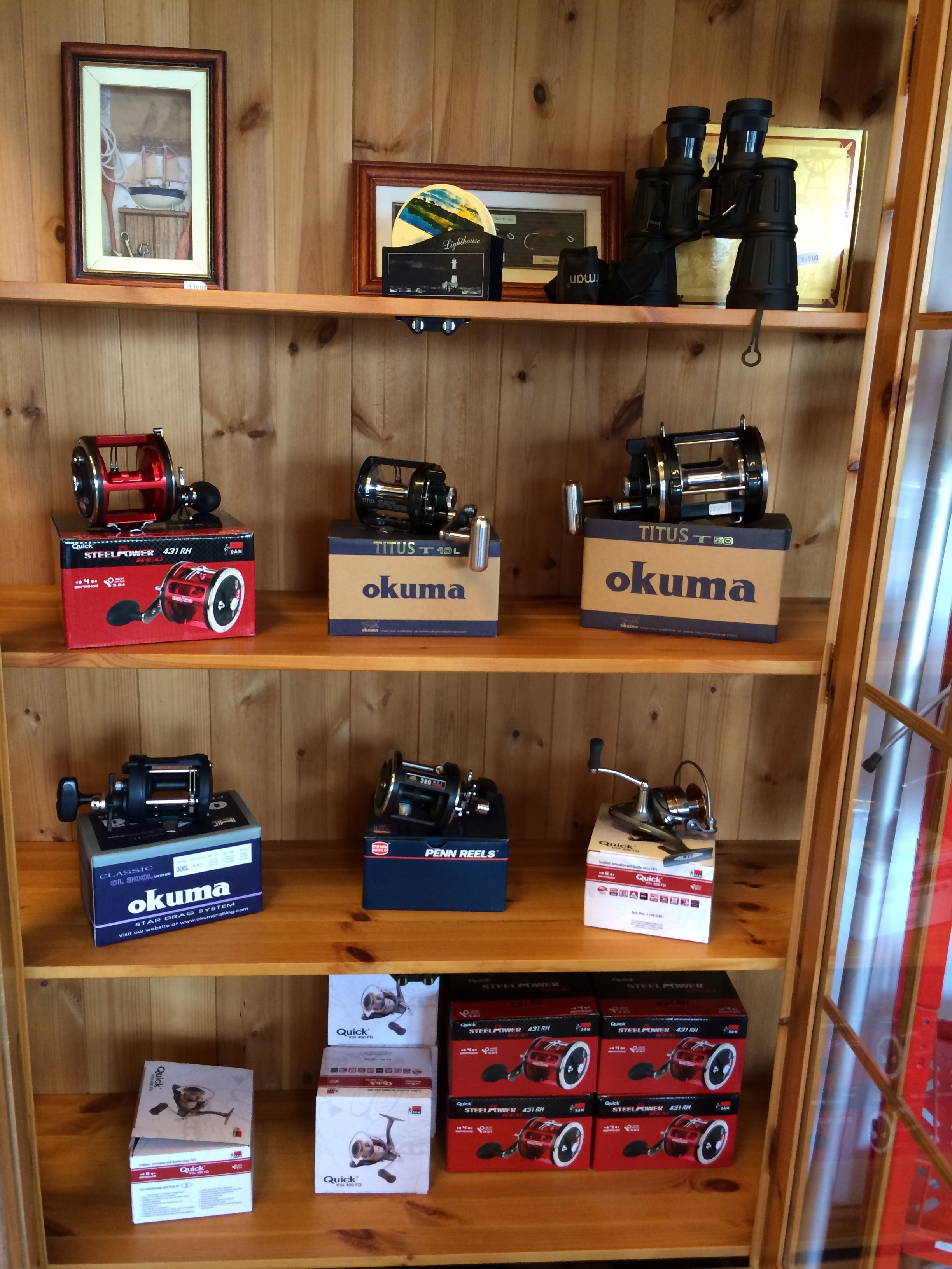 Un estante de madera con varios carretes y accesorios de pesca, incluyendo marcas como Okuma y Penn Reels.