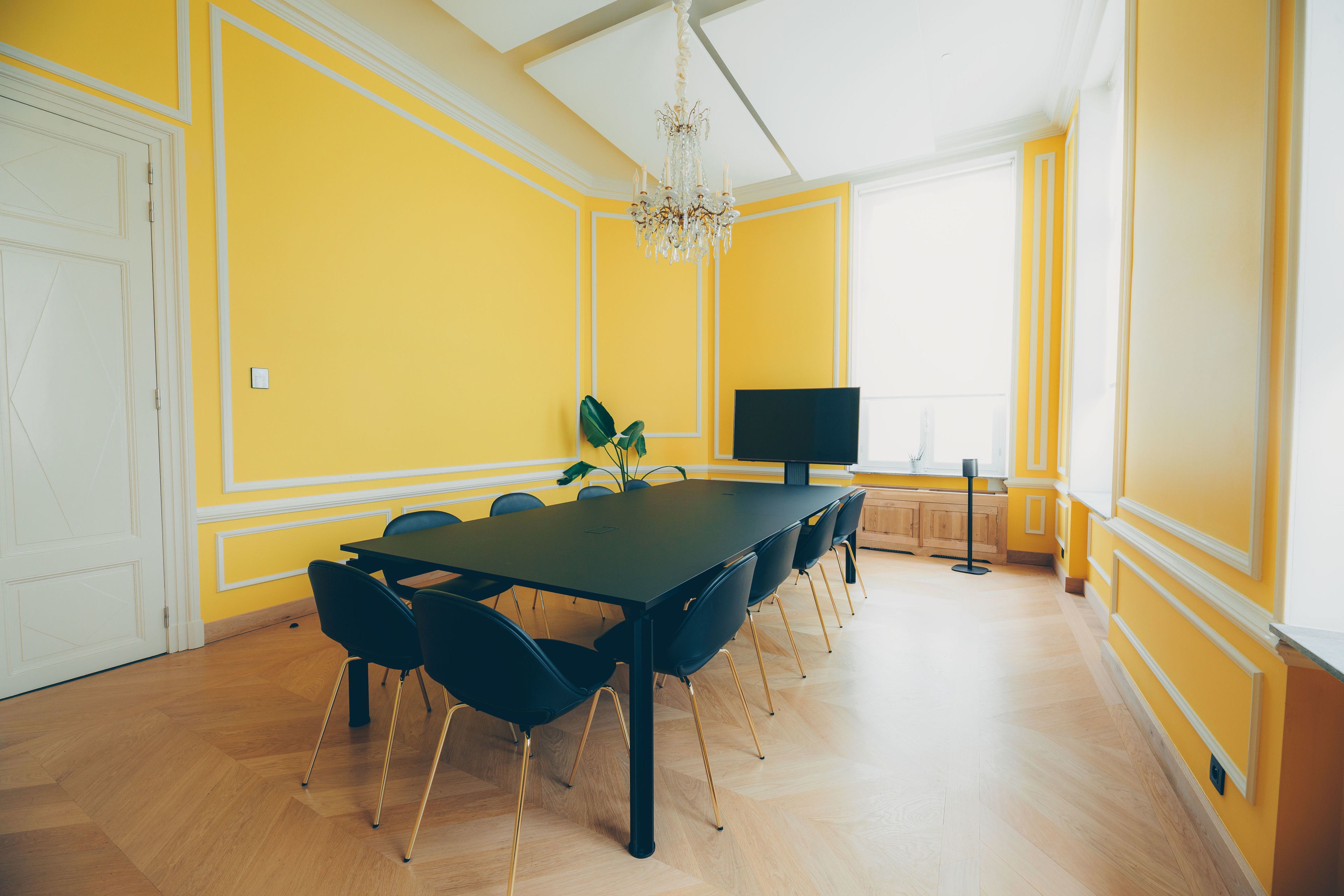 A dining area with a long table, black chairs, a chandelier, and yellow walls.