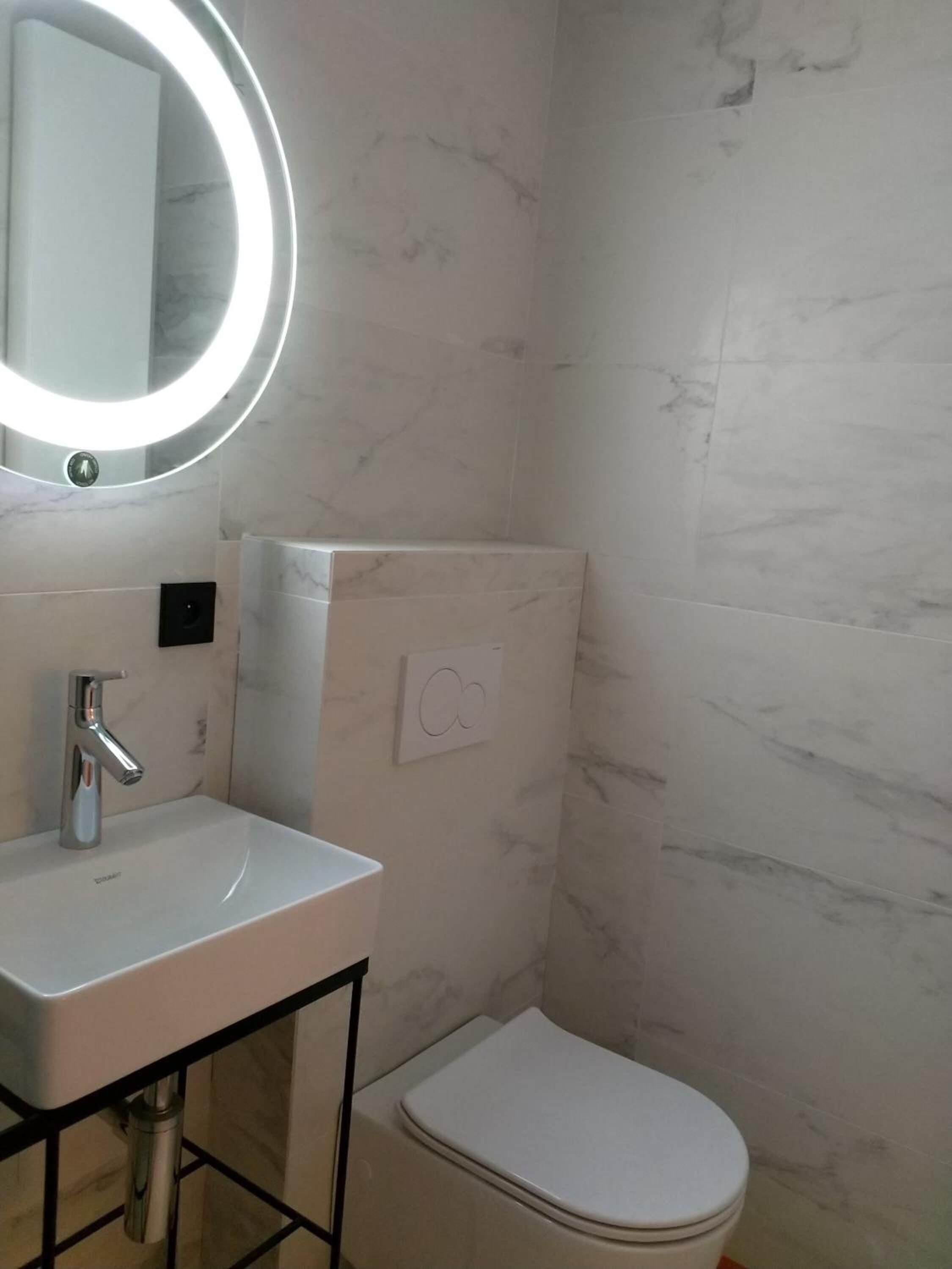 Un baño con un lavamanos blanco, un espejo con luz, un inodoro y una pared de mármol.