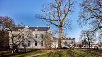 A large, historic building with a symmetrical facade, a prominent central tree, and a well-maintained lawn.
