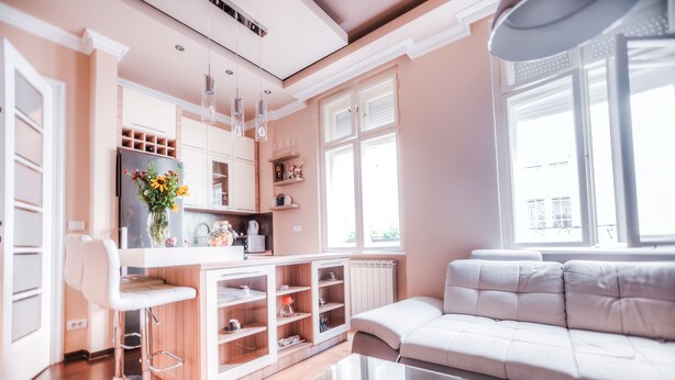 A modern kitchen with a white refrigerator, wooden cabinets, and a bar stool. There is a glass coffee table and a white sofa in the living area.