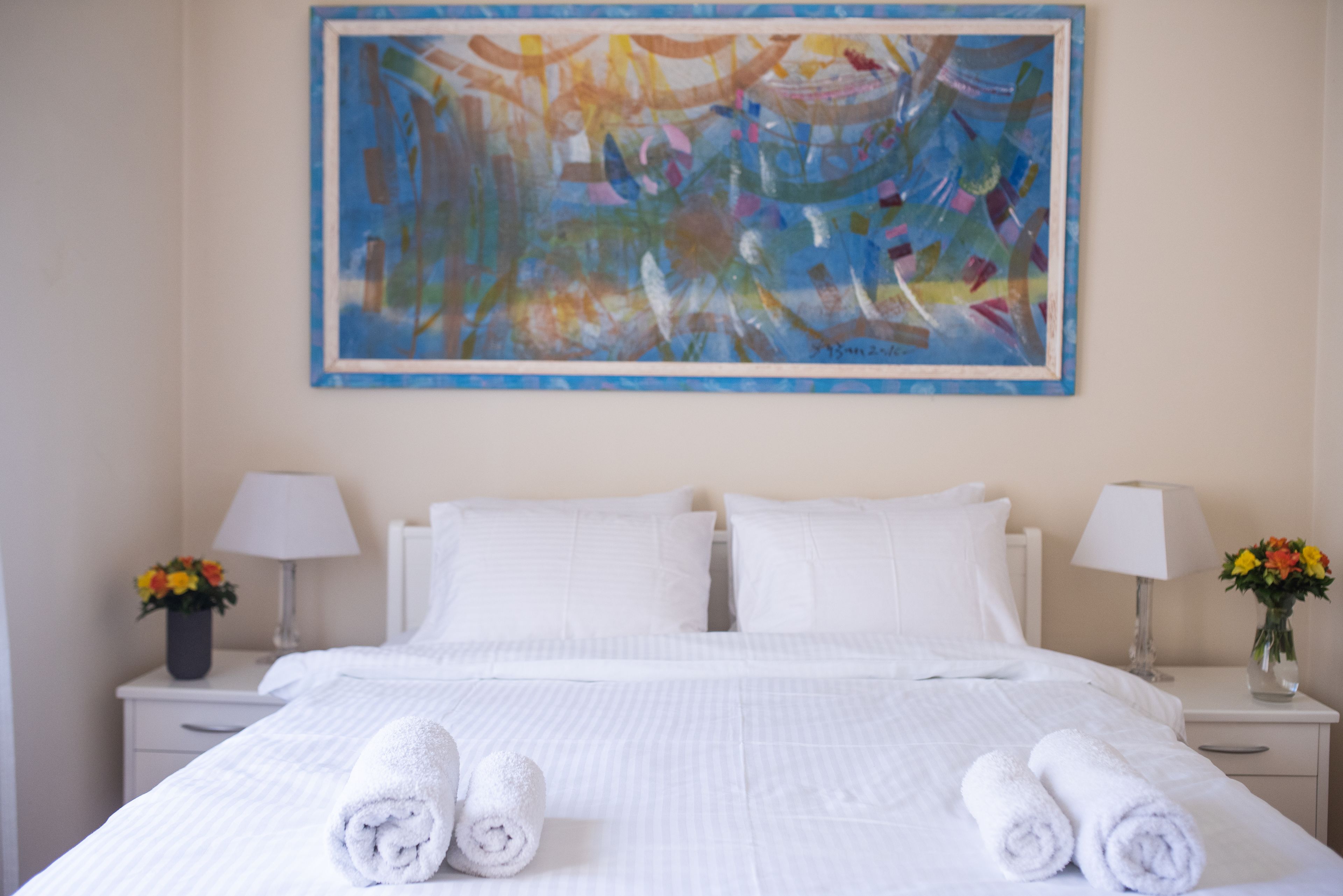 Une chambre d’hôtel avec un grand tableau abstrait au mur, un lit soigneusement fait avec du linge de maison blanc, deux tables de chevet avec des lampes et des vases à fleurs.