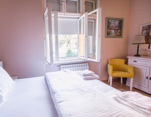Une chambre à coucher avec un lit, une fenêtre dont les volets sont ouverts, un radiateur, une chaise jaune et une commode blanche.