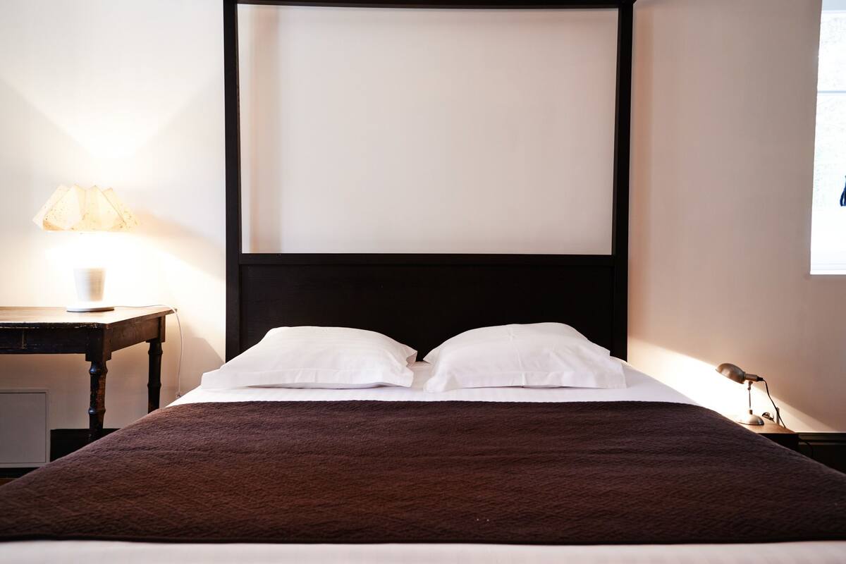 Un lit bien fait, avec des oreillers blancs et une couverture marron, une table de chevet avec une lampe, et une fenêtre avec des stores.