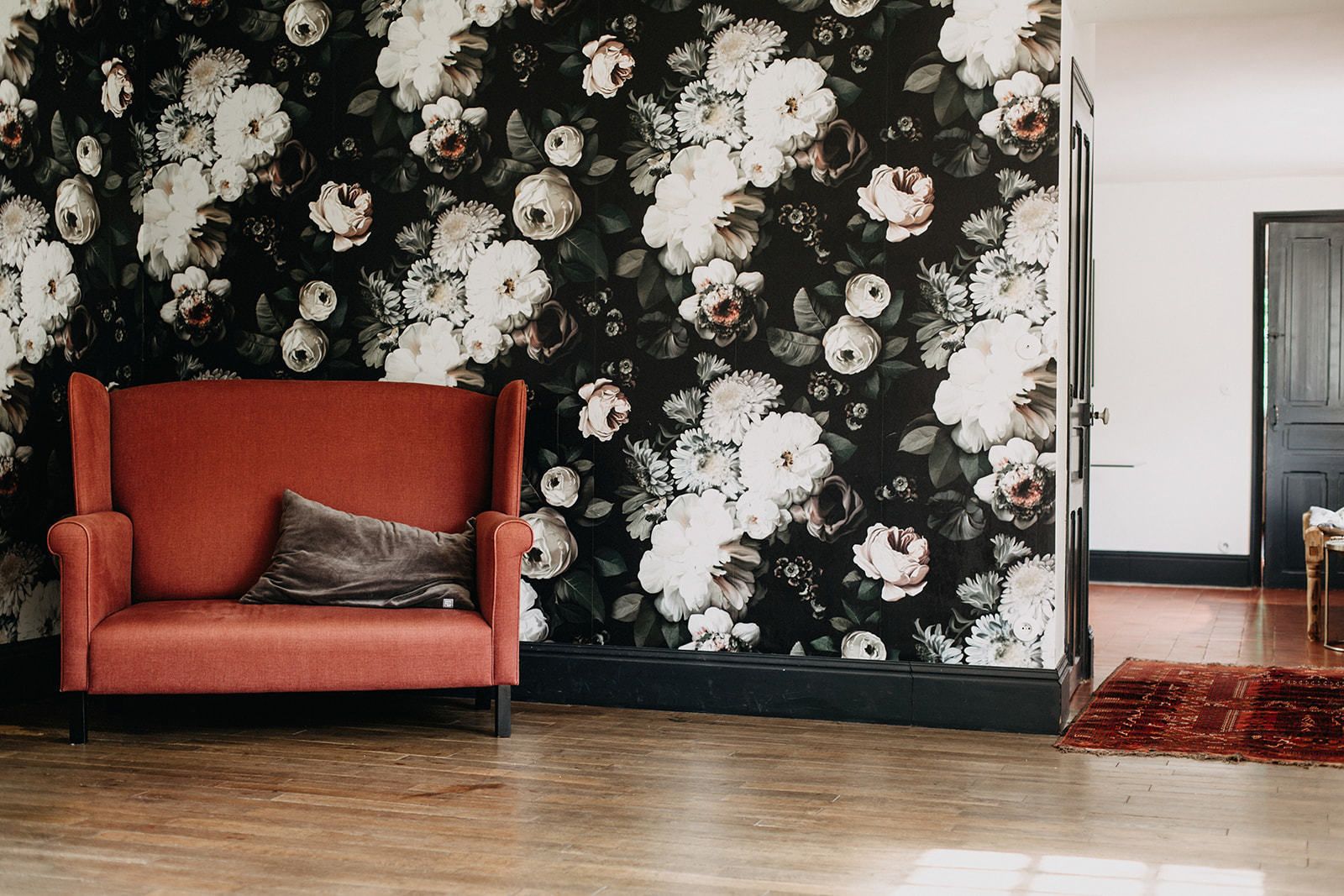 Una habitación con papel tapiz floral oscuro, un sillón rojo con cojín y suelo de madera.