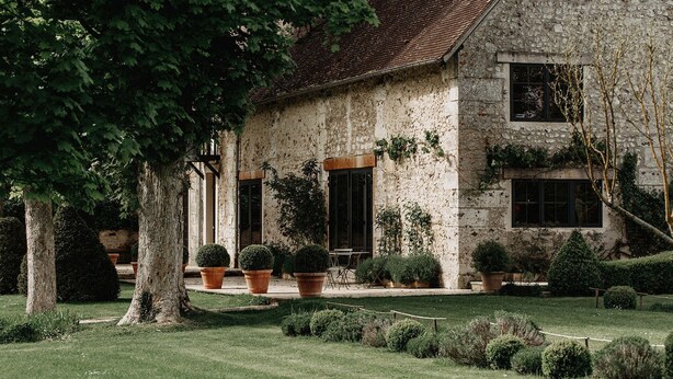 A stone building with a tiled roof, surrounded by a well-maintained garden with various potted plants and trimmed hedges. - La Dime de Giverny - Chambres D'hôtes (Giverny)
