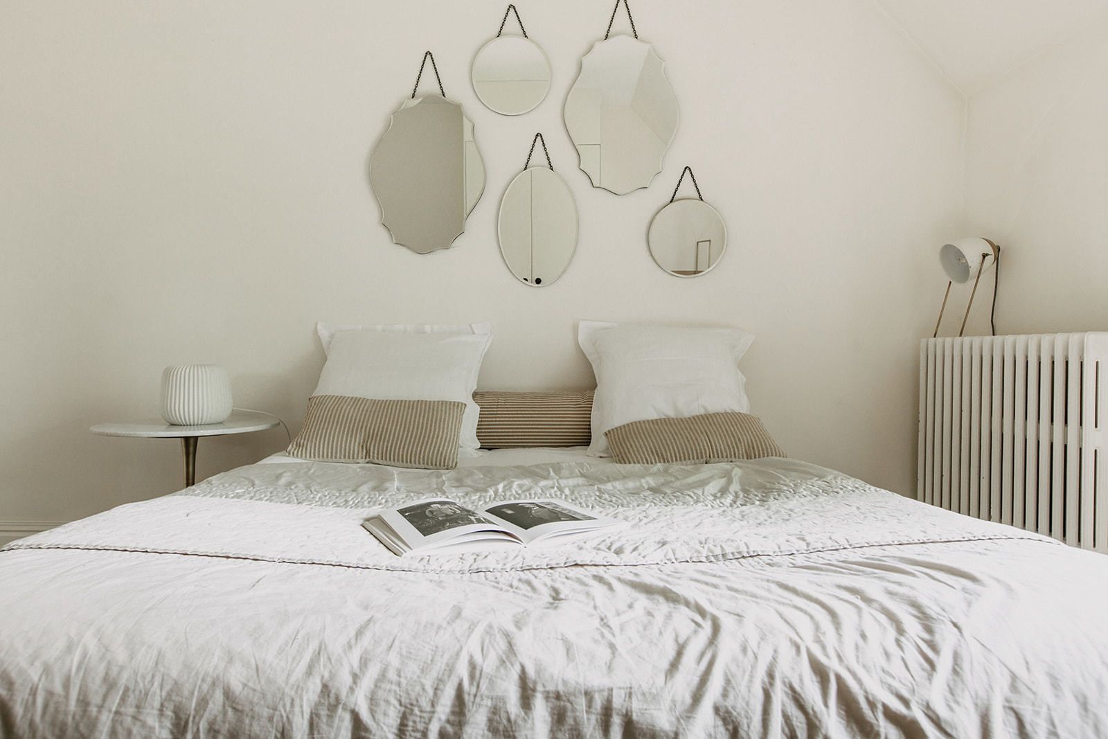 Un lit bien fait, avec un livre posé dessus, une table de chevet avec une lampe et un radiateur au mur.