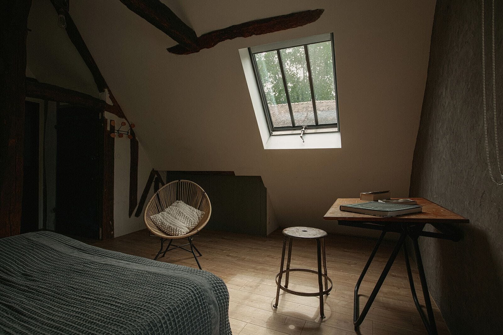 A cozy attic bedroom with a sloped ceiling, a large window, a wooden table, a wicker chair, and a bed with a textured blanket.