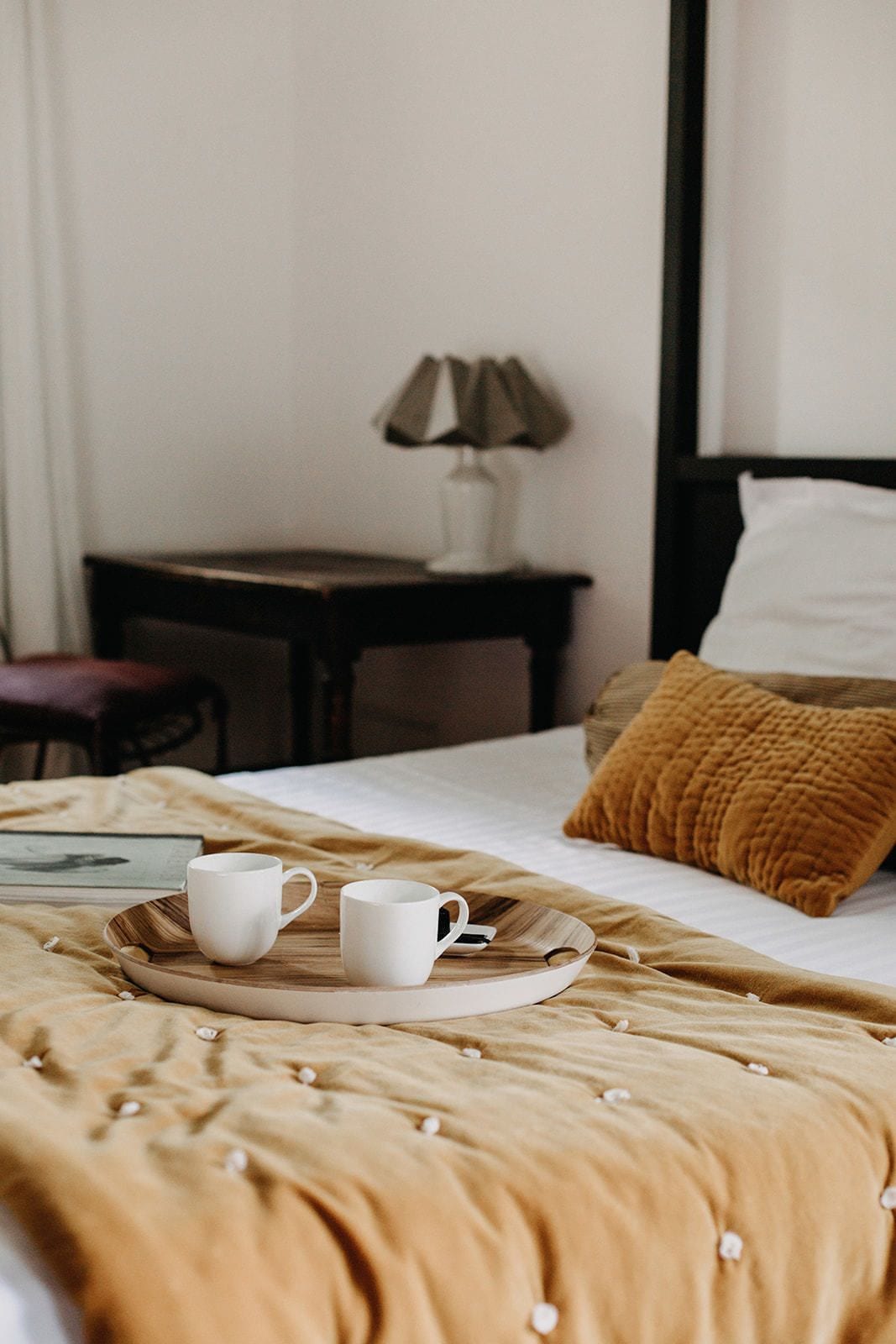 Una cama bien hecha con una manta marrón y almohadas, una mesita de noche con una lámpara, y dos tazas blancas sobre una bandeja.