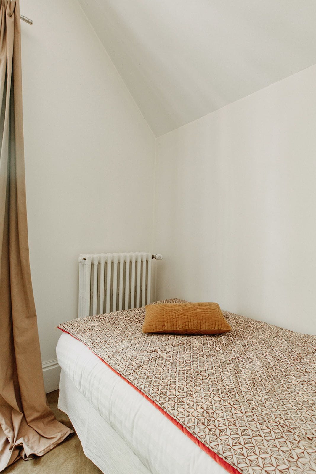 Quarto pequeno com cama de solteiro, colcha estampada, radiador e cortina.