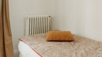 Quarto pequeno com cama de solteiro, colcha estampada, radiador e cortina.