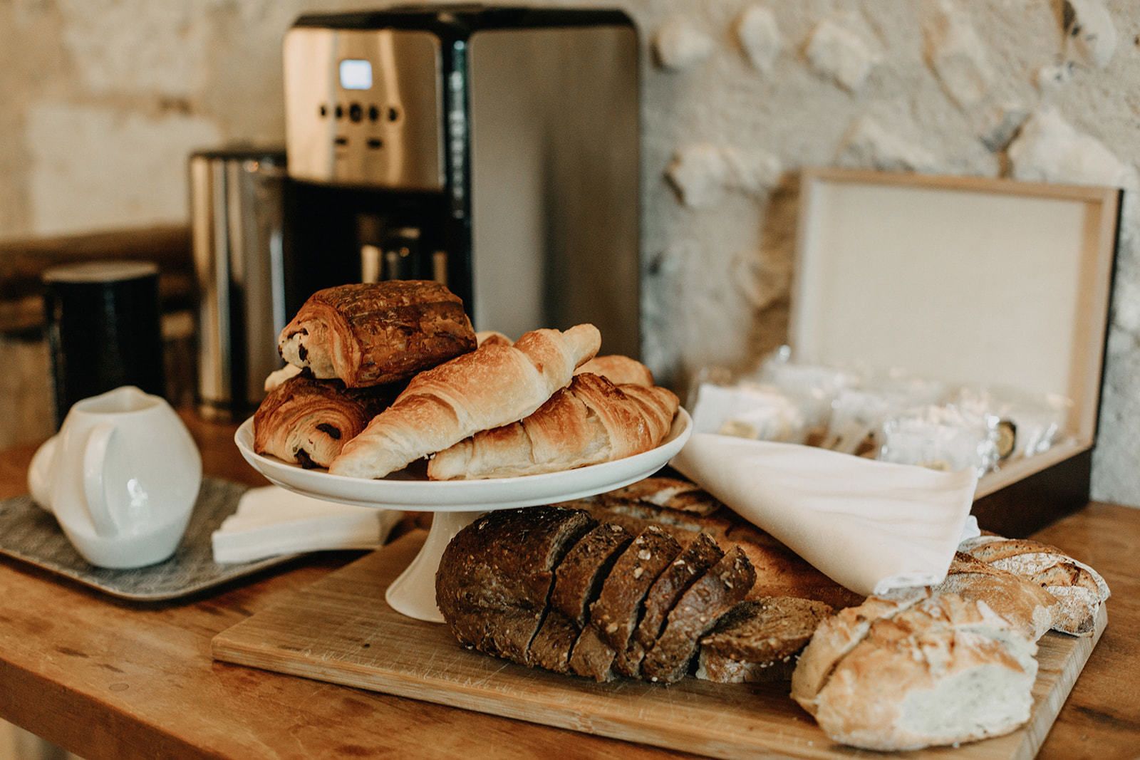 A wooden table with a variety of breads and pastries, a coffee machine, and a white teapot.