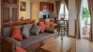 A cozy living area with a sofa, coffee table, and a dining table with chairs. There is a kitchen area with wooden cabinets and a window with curtains. - The Lighthouse (Can Tho)