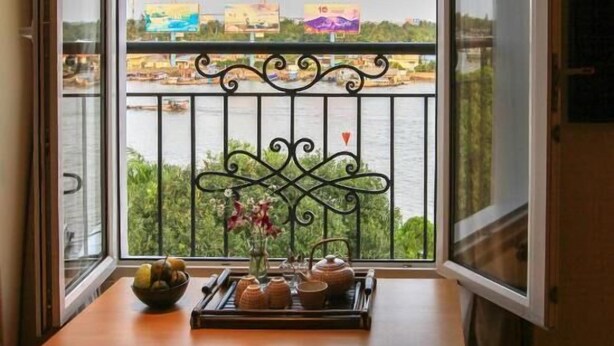 A room with a view of a river and buildings across it, a small table with a tray of items, and a chair with a red cushion. - The Lighthouse (Can Tho)