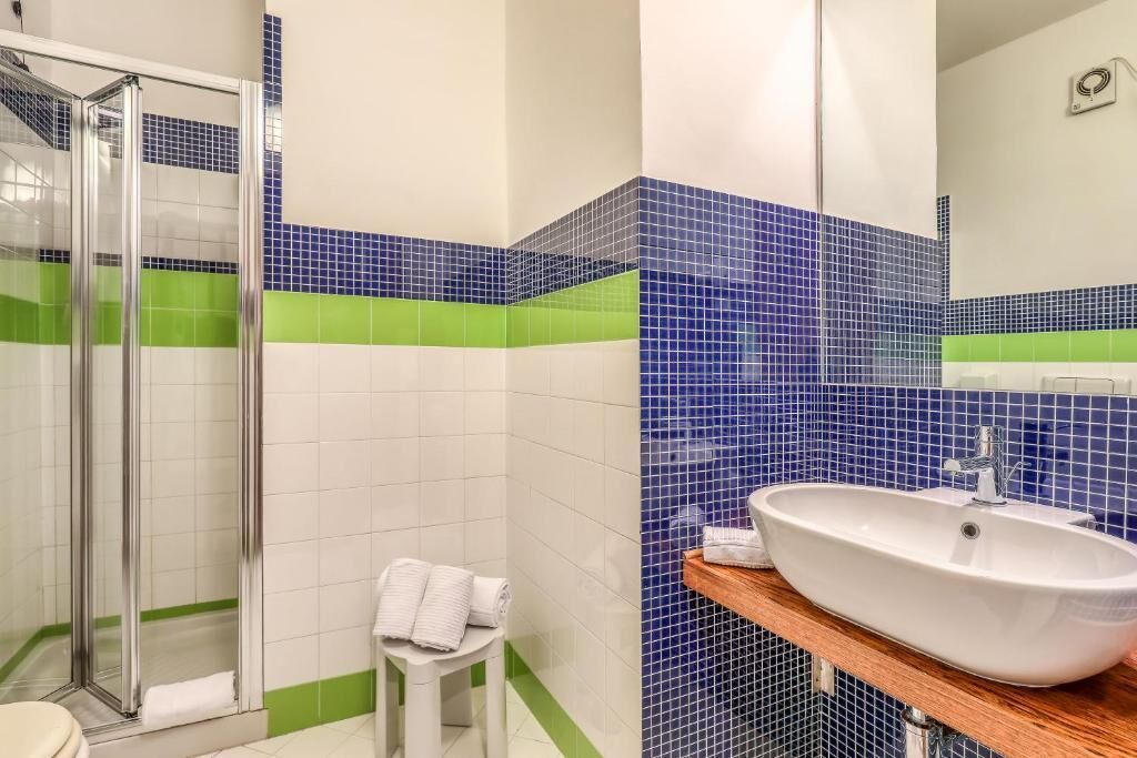 A bathroom with a glass shower enclosure, a white sink with a wooden countertop, and a green and blue tiled backsplash.