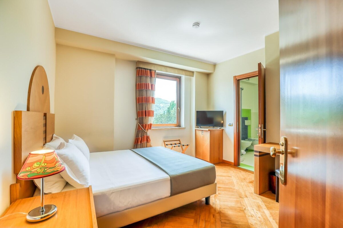 Habitación de hotel con cama, escritorio con lámpara, televisión y vistas al exterior.