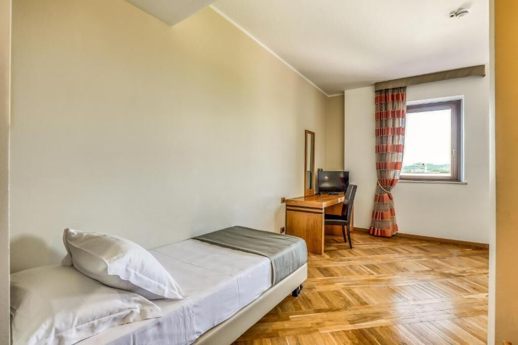 A small, single-bed room with a wooden desk and chair, a television, and a window with curtains.