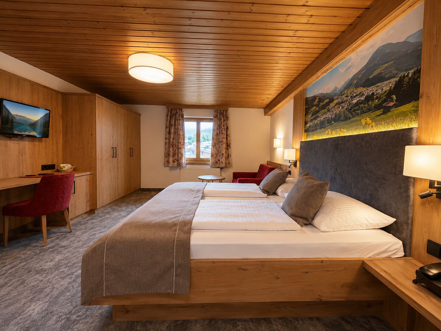 A hotel room with a wooden ceiling, a bed with two pillows, a desk with a chair, a television, and a painting of a landscape.