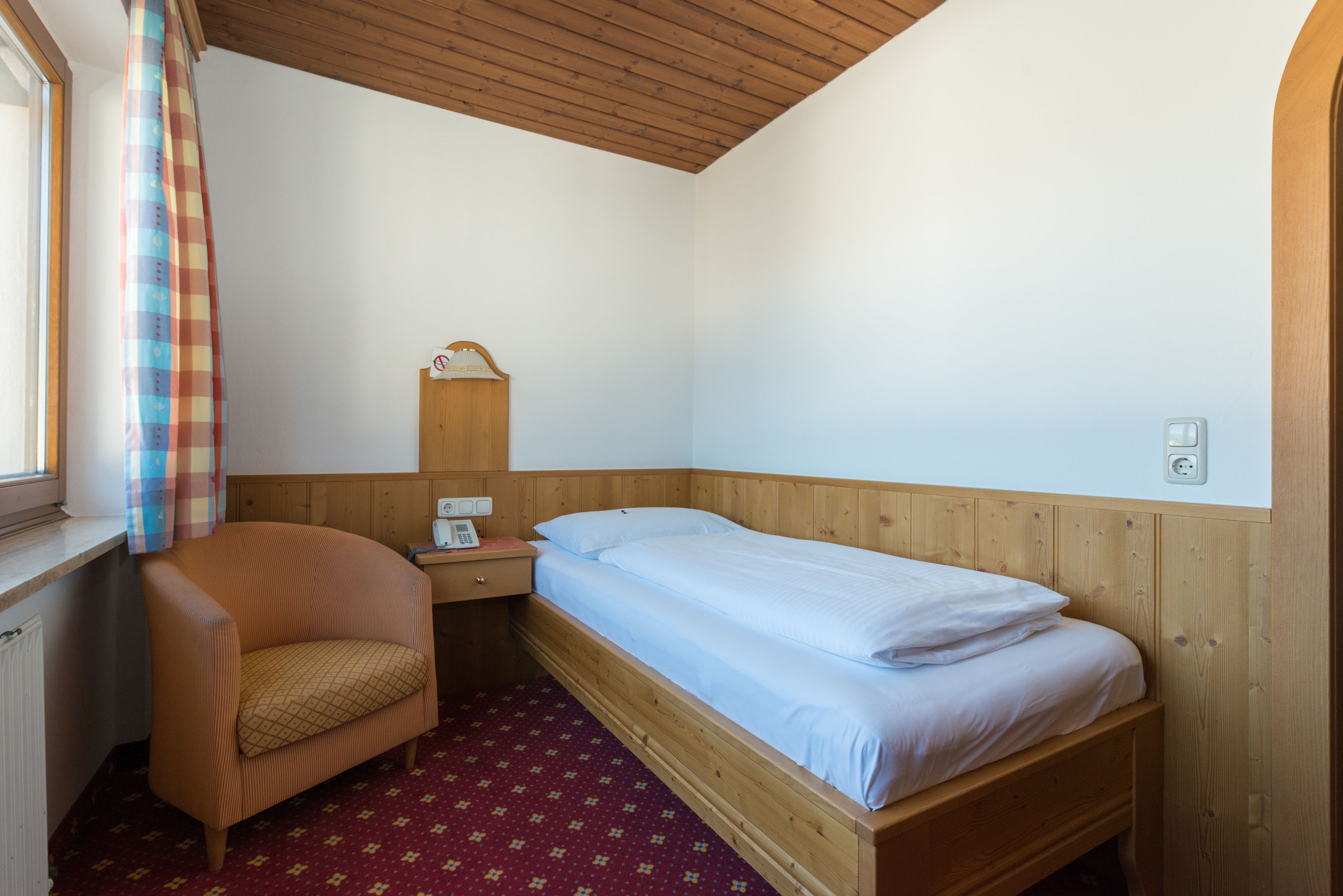 A small, neatly arranged bedroom with a single bed, a chair, a nightstand, and a wooden headboard.