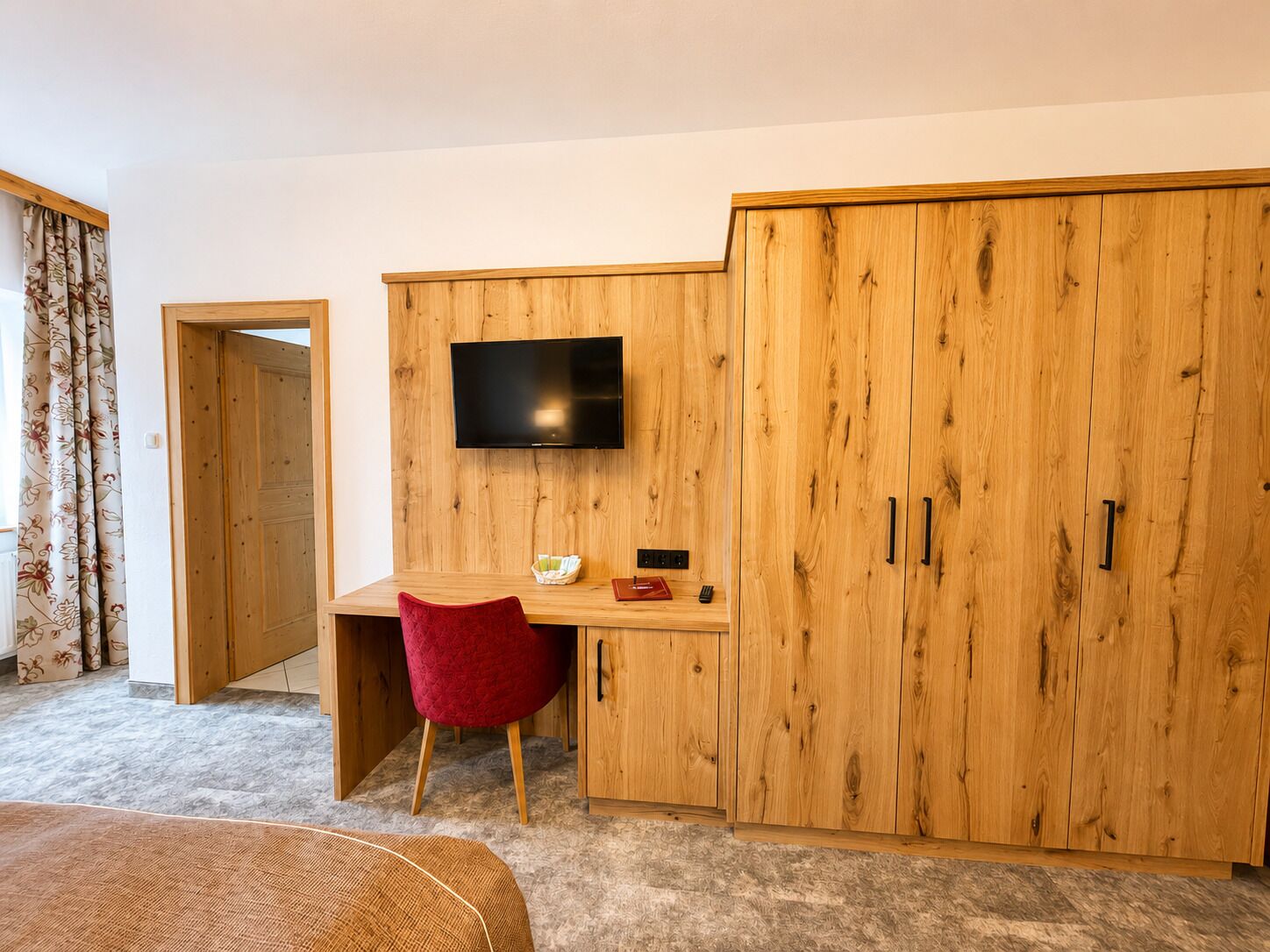 A room with a wooden wardrobe, a desk with a chair, a television mounted above the desk, and a bed in the foreground.