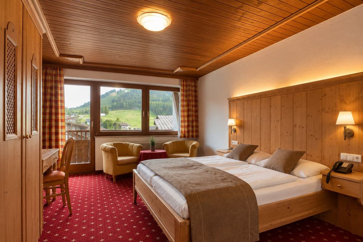 A room with a bed, two chairs, a desk, a window with a view of the countryside, and a wooden ceiling.