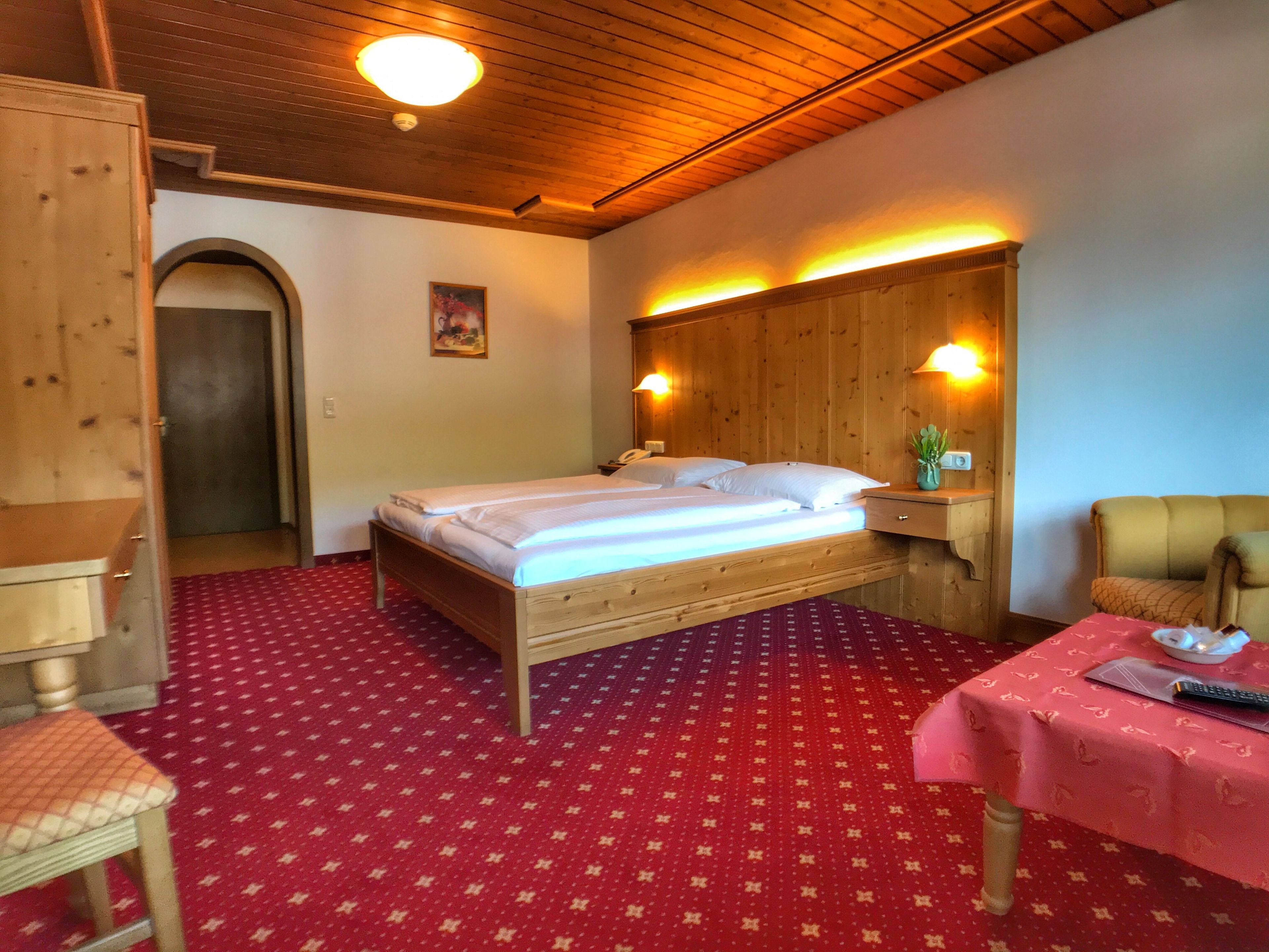 A wooden bed with white linens in a room with a red carpet, a wooden wardrobe, a desk with a plant, and a chair.
