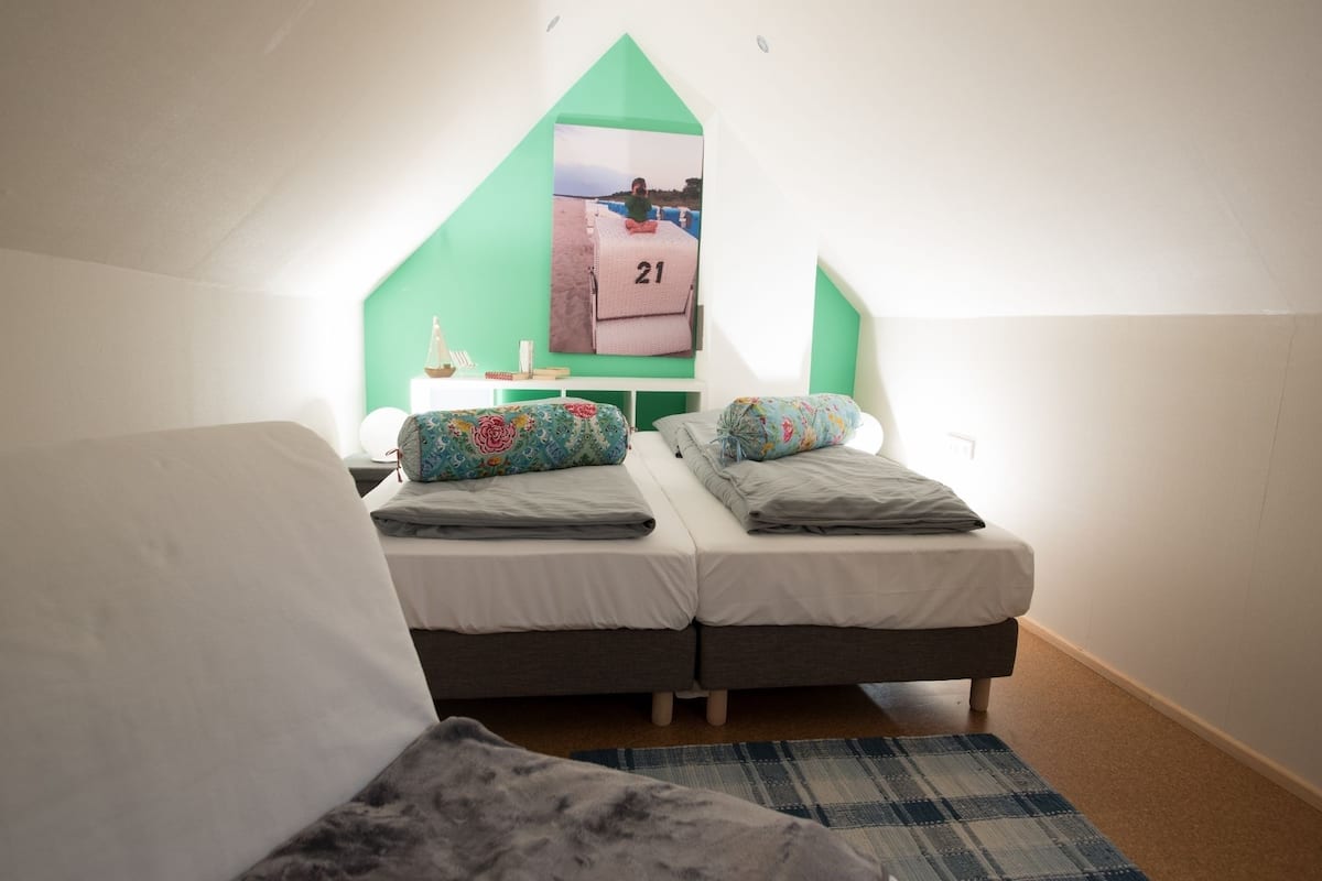 A small, cozy attic bedroom with two beds, a green wall, and a window overlooking a scenic view.