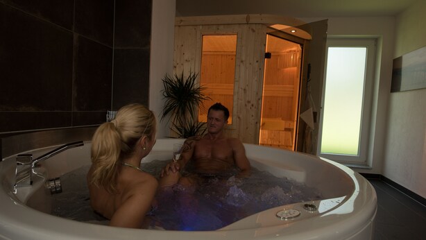 A couple in a hot tub with a wooden interior and a large window. - Seeland Lodge (Hilpoltstein)
