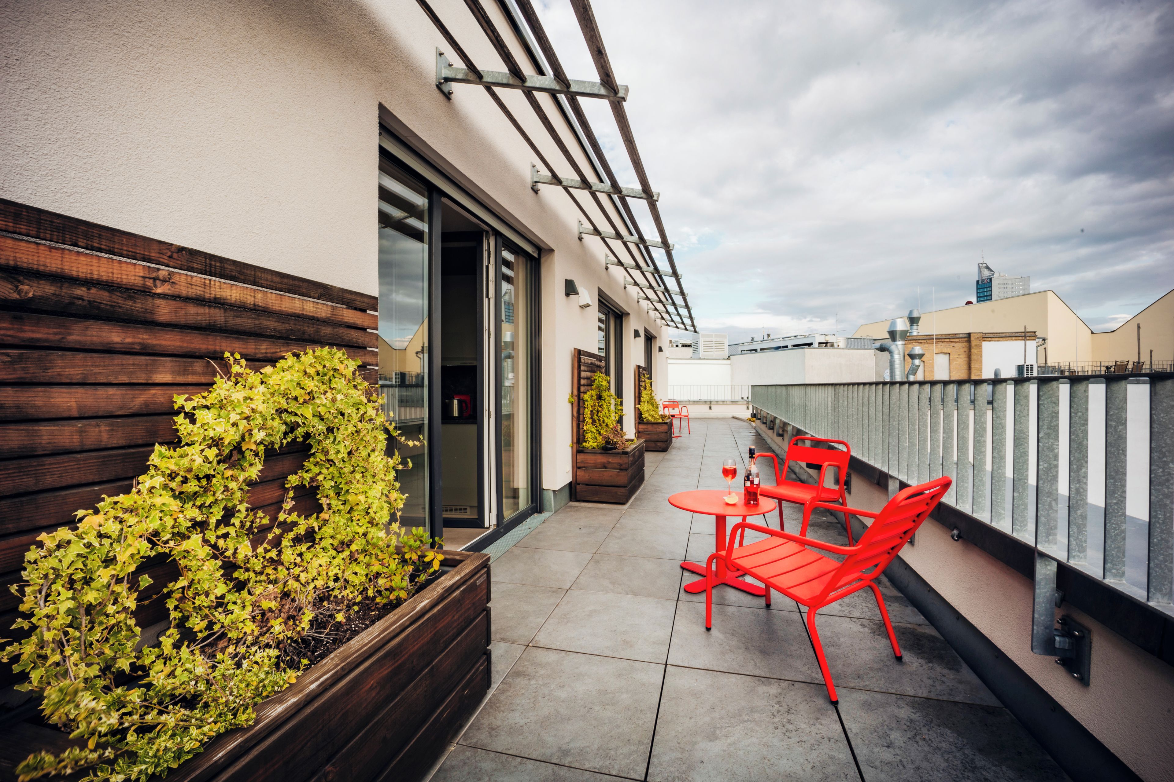 A rooftop terrace with a wooden planter, red chairs, and a glass door leading to an interior space.