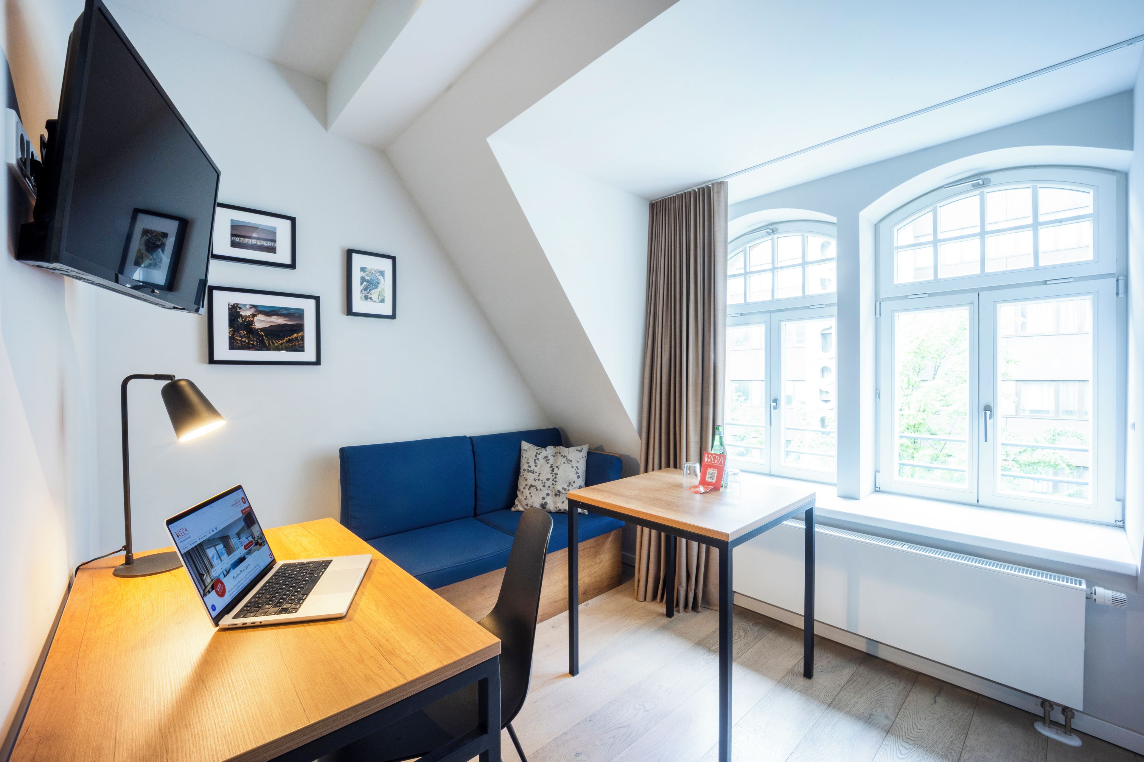 Ein modernes Zimmer mit einem Laptop auf einem Holztisch, einem blauen Sofa, einem kleinen Tisch mit einer Flasche und einem Fenster mit Vorhängen.
