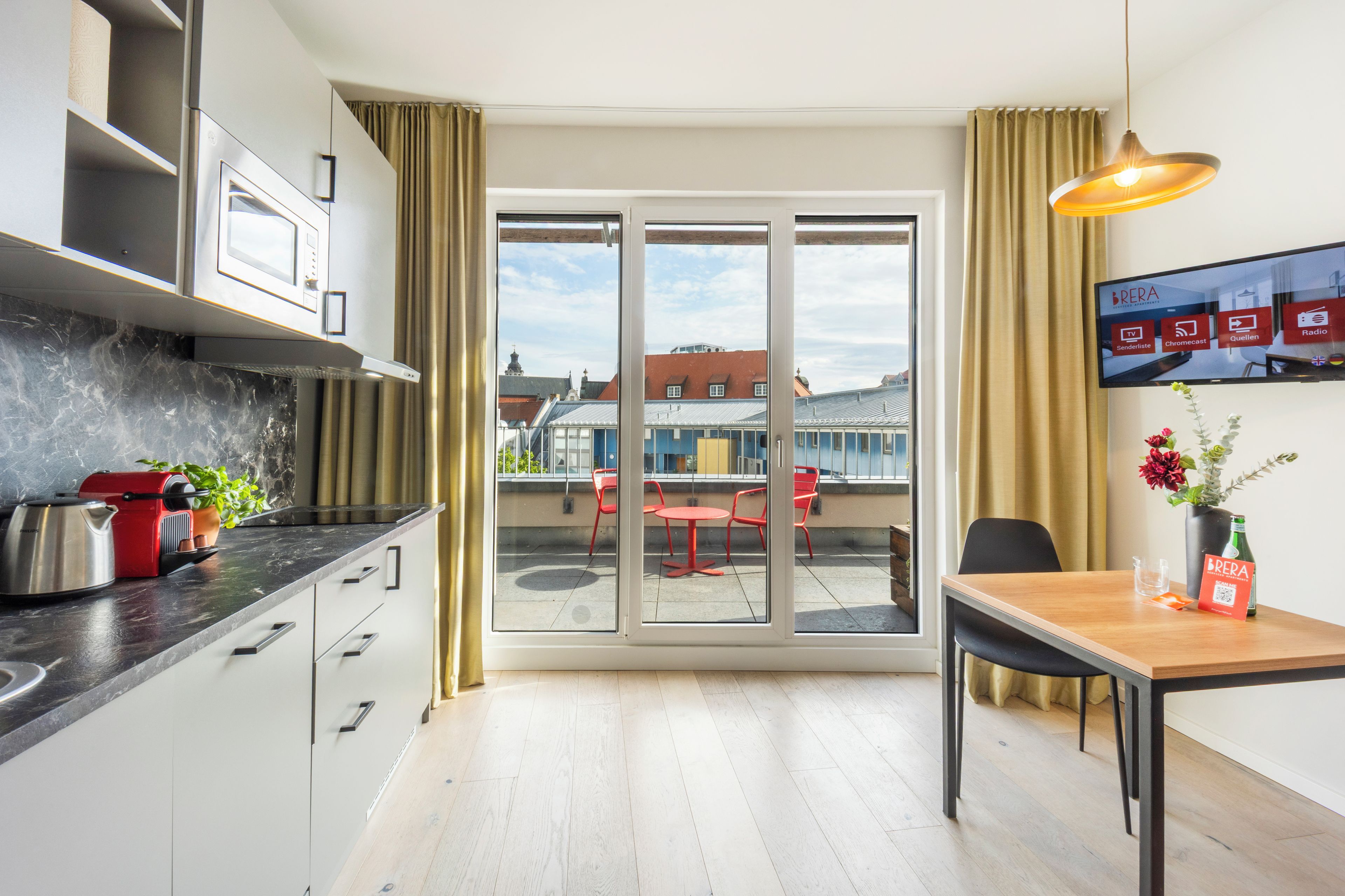 A modern kitchen with a dining area, a view of the cityscape through a glass door, and a table with a vase of flowers.