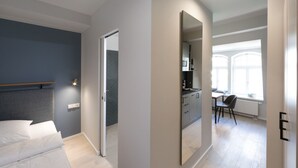 A modern bedroom with a bed, a bedside table, and a wardrobe. There is a bathroom with a shower and a toilet. The room has a large window with natural light. - Brera Serviced Apartments Leipzig (Leipzig)