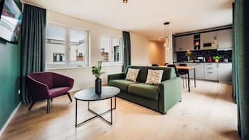 Een moderne woonkamer met een groene bank, een rode fauteuil en een kleine ronde tafel. Er is een keukenhoek met een eettafel en stoelen.