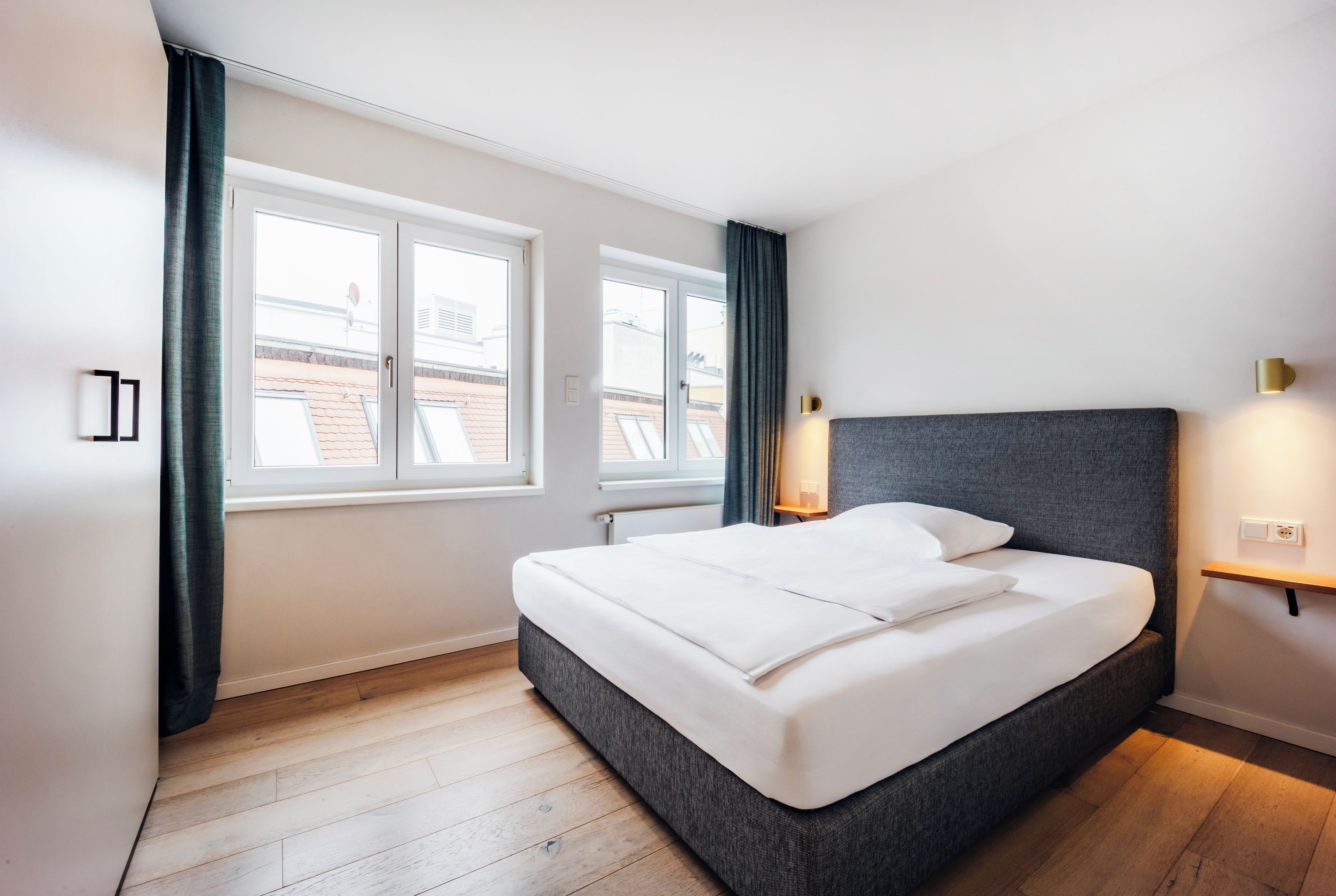 Ein modernes Schlafzimmer mit einem großen Bett, zwei Fenstern, einem Nachttisch und einer Wandleuchte.