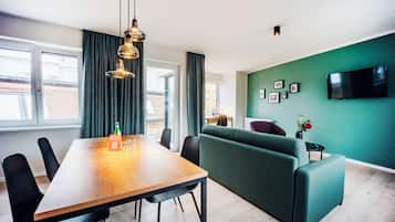 A modern living room with a wooden dining table, green sofa, and a flat-screen TV mounted on the wall.