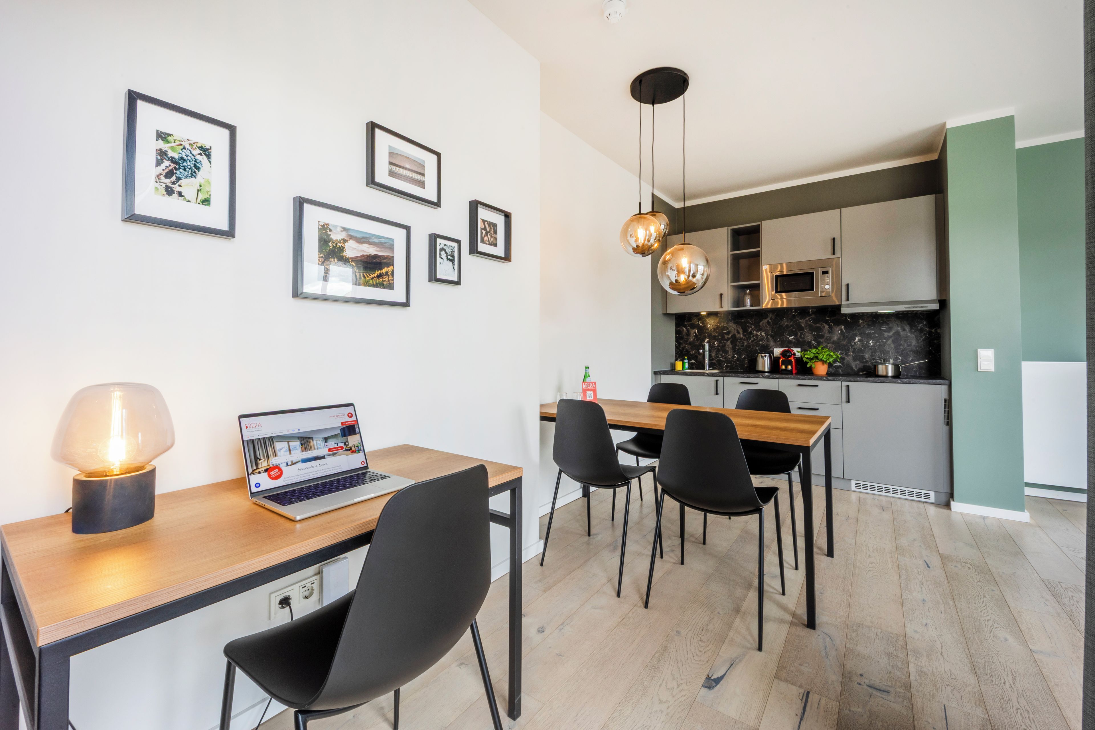 A modern kitchen with a dining area, featuring a laptop on a table, framed pictures on the wall, and pendant lighting.
