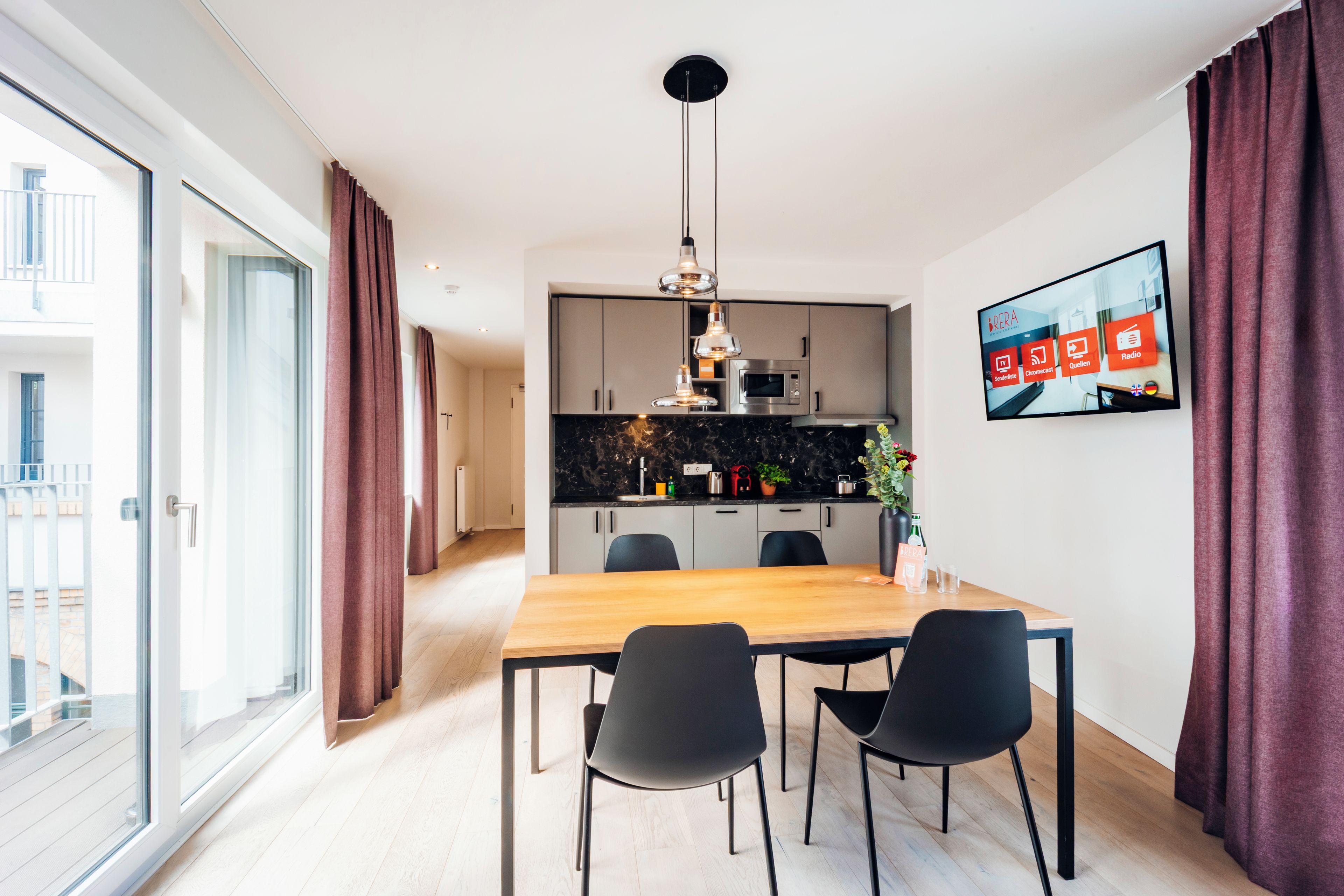 A modern kitchen with a dining area, a TV mounted on the wall, and a sliding glass door leading to an outdoor balcony.